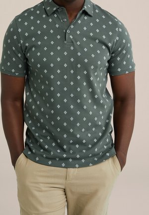 Uomo che indossa una polo verde a maniche corte con forme a rombo bianche e pantaloni beige, con le mani nelle tasche, su uno sfondo semplice.