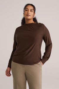 Top marrone a maniche lunghe con scollo alto, dettaglio arricciato e bottoni dorati sulla spalla. Abbinato a pantaloni su misura color beige chiaro.