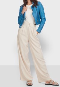 Una chaqueta de motocicleta de cuero azul, una blusa blanca, pantalones anchos beige y sandalias trenzadas grises crean un conjunto casual en capas.