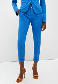 Blå skräddarsydda byxor med en slät yta, har en slim fit och rullade fållar. Kombineras med orange, remförsedda högklackade skor.