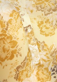 Blusa floreale in morbido tessuto giallo chiaro, con grandi motivi floreali marroni e dorati, e tre bottoni bianchi sulla parte anteriore.