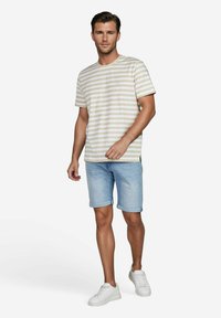 Gestreiftes Baumwoll-T-Shirt in Beige und Creme, kombiniert mit hellblauen Jeansshorts. Das Model trägt weiße Sneaker und steht vor einem schlichten Hintergrund.