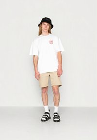 Weißes übergroßes T-Shirt mit rotem Grafikdruck, beigefarbene Shorts, schwarzer Fischerhut, weiße Socken und schwarze Sandalen mit weißen Riemen.