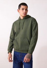 Grüne Baumwollkapuzenjacke mit einer Fronttasche, Kordeln und gerippten Bündchen, kombiniert mit blauen Jeans, präsentiert auf einem Model vor einem hellen Hintergrund.