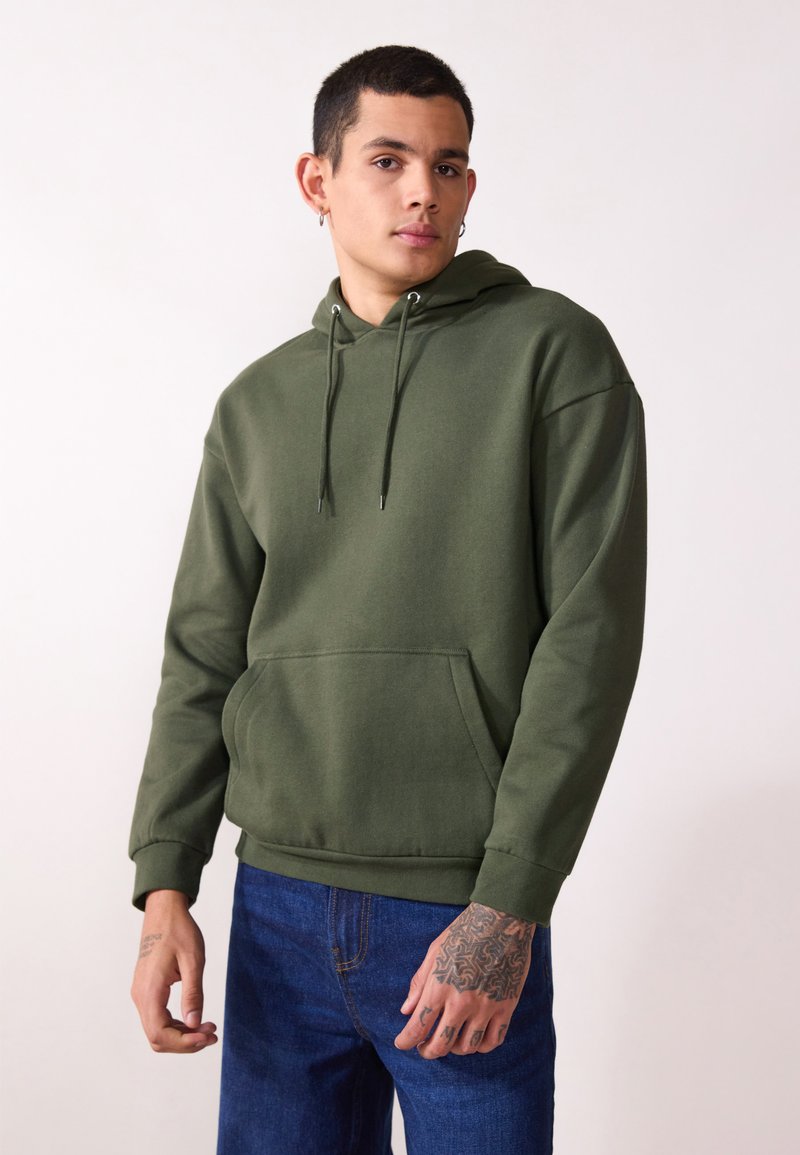 Grønn bomull hettegenser med en frontlomme, snor, og ribbestrikkede cuffs, sammen med blå jeans, vist på en modell mot en lys bakgrunn.