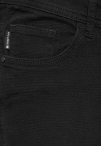 Schwarze Jeans aus Stretchstoff, mit einer Vordertasche und einem Etikett. Die Textur ist glatt, mit tonalem Nähen und einem Slim-Fit-Design.