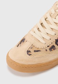 Sneaker en suède beige avec un accent en imprimé léopard, des lacets plats crème et une semelle en gomme texturée avec un motif en zigzag sur le contour.