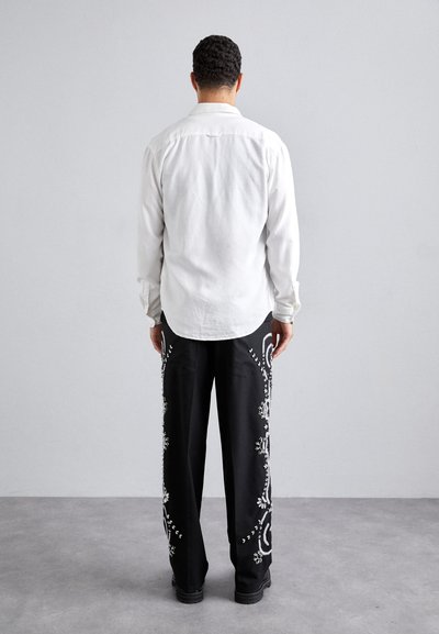 Fehér, hosszú ujjú ing, amelyet fekete, bő lábú nadrággal párosítanak, amelyet részletes fehér virágminták díszítenek az oldalán.