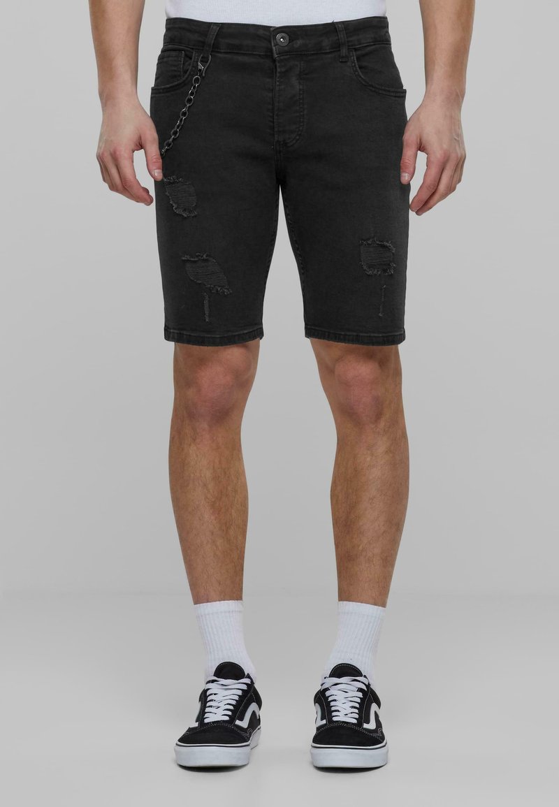 Shorts in denim nero con dettagli strappati, passanti per cintura e una catena laterale. Taglio aderente, che arriva poco sopra le ginocchia, abbinati a sneaker nere.