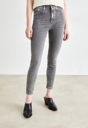 Vrouw die een grijze skinny jeans met hoge taille en zwarte muiltjes met hakken draagt, staand op een lichte vloer tegen een witte muur.