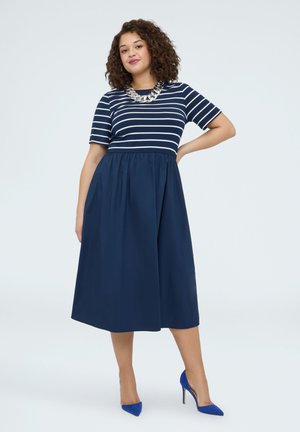 Frau mit lockigem Haar, die ein marineblaues Kleid mit weißen Streifen oben, eine silberne Halskette und blaue High Heels trägt, steht mit einer Hand in der Hüfte.