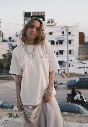 Kremowy, oversizowy t-shirt z krótkimi rękawami i subtelnym haftem „Surfin'”. Do tego jasnobeżowa, jedwabista spódnica. Srebrne łańcuszkowe dodatki.