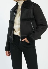 Chaqueta bomber de ante negra con acentos de piel de oveja, bolsillos con cremallera y cuello alzado, con un diseño corto y puños texturizados.