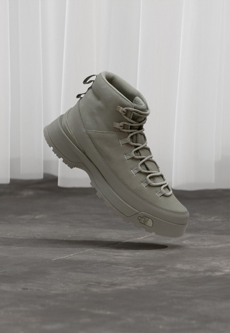 Ανοιχτό γκρι ψηλό μποτάκι πεζοπορίας με κορδόνια και λογότυπο The North Face στη σόλα, που φαίνεται να αιωρείται πάνω από τσιμεντένιο δάπεδο μπροστά από λευκές κουρτίνες.