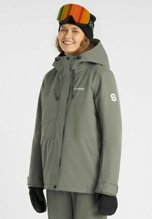 Smilende person iført olivengrøn vinterjakke, sorte handsker, sort hue og orangefarvede skibriller mod ensfarvet baggrund.