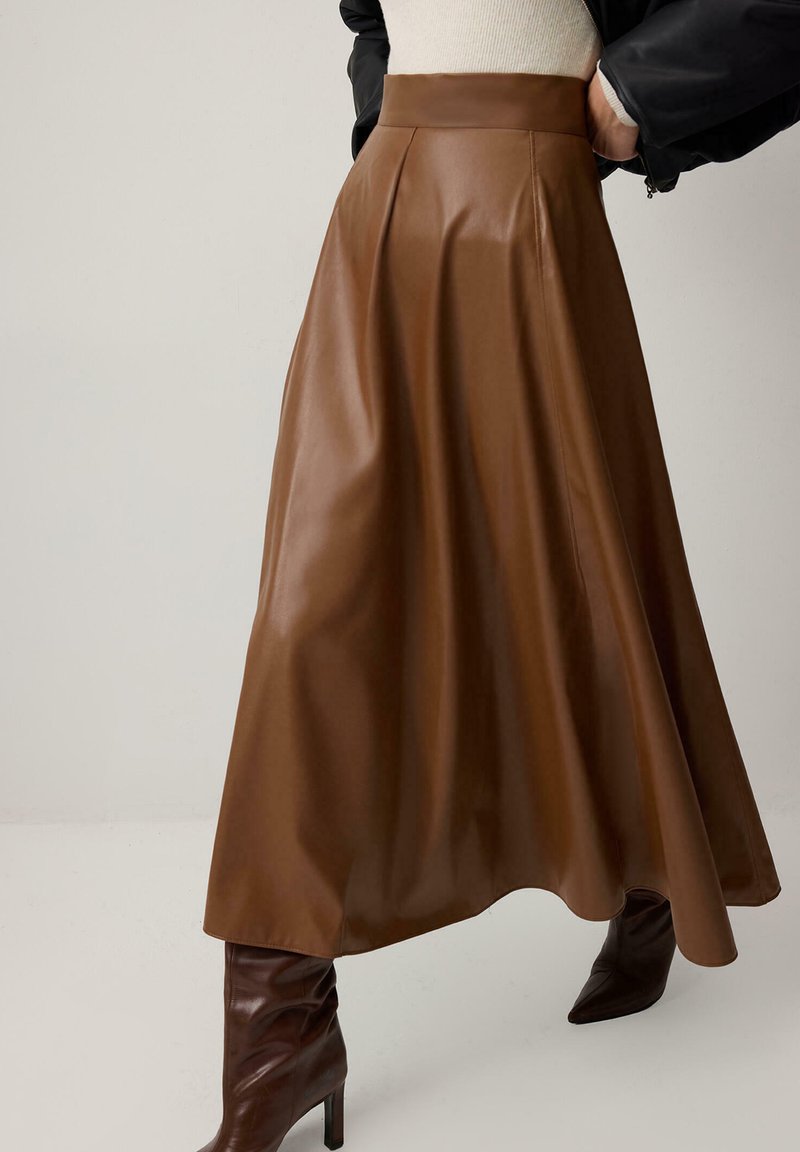 Καφέ midi φούστα από ψεύτικο δέρμα με ψηλή μέση, συνδυασμένη με καφέ σκούρα μποτάκια με τακούνι, φορεμένη με ανοιχτόχρωμο τοπ και μαύρο μπουφάν.