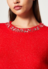 Rode trui met een gestructureerd breipatroon en een collier versierd met heldere en zilveren sieraden.