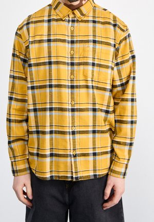 Mężczyzna ubrany w żółtą, czarno-białą koszulę w kratę z kieszenią na piersi oraz ciemne spodnie na jednolitym tle.