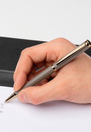 Grau gefärbter Stift mit goldenen Akzenten, gehalten von einer Hand, die auf weißem Papier schreibt. Der Hintergrund zeigt eine schwarze, strukturierte Oberfläche.