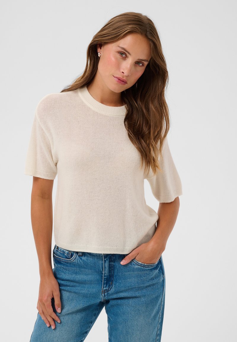 Femme aux longs cheveux bruns, portant un haut en maille crème à manches courtes et un jean bleu, debout avec une main dans la poche sur fond blanc.