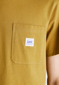 T-shirt giallo senape in morbido cotone. Dotato di una tasca sul petto con un'etichetta bianca e viola con la scritta "Lee WORKWEAR". Texture liscia.