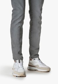 Baskets blanches et beige avec une tige en maille, des accents en daim, et une semelle en caoutchouc texturée, associées à un pantalon gris slim.