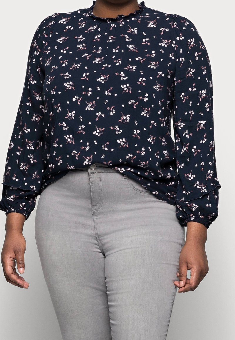 Marinblus med blommönster och volangkrage, långa ärmar med elastiska manschetter, prydd av små vita och rosa blommor. Grå byxor syns.