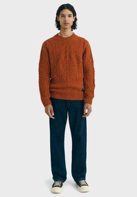 Jeune personne avec des cheveux bouclés portant un pull texturé couleur rouille, un jean bleu foncé et des baskets noir et blanc, se tenant face à l'avant.