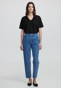 T-shirt nera a V, vestibilità comoda, abbinata a jeans blu a gamba dritta a vita alta. La modella indossa scarpe nere senza tacco e accessori minimalisti, su uno sfondo neutro.