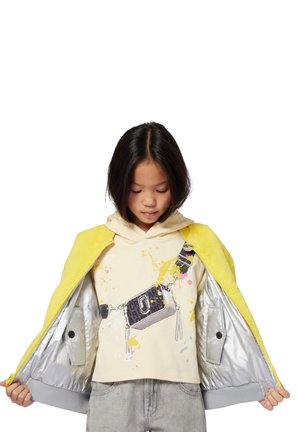 Geel donzige jacket over een beige hoodie met een grafische print; grijze binnenkant met een soepele textuur; grijze jeans en een hand die het jacket vasthoudt.
