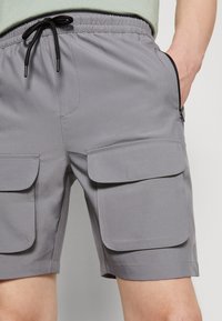 Šedé cargo šortky s elastickým pasem a šňůrkou. Mají dvě velké přední kapsy s klopovými uzávěry a hladkou, texturovanou tkaninu.