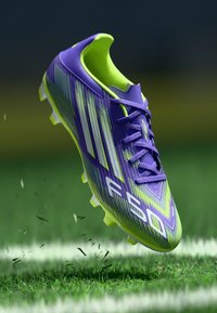 Crampons de football violet et vert lime avec un design épuré, comportant trois bandes blanches, une surface texturée et des crampons jaunes.