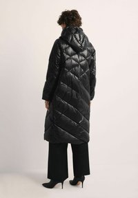 Czarna kurtka puchowa o skośnym pikowanym wzorze, z kapturem i długości do połowy uda, zestawiona z szerokimi czarnymi spodniami i butami na obcasie.