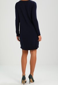 Robe en tricot marine à manches longues, coupe au genou et col rond. Associée à des chaussures à talons verts. Texture lisse.