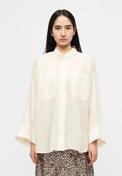 Ελαφρώς μπεζ πουκάμισο με κουμπιά και μανδρινό γιακά, με κάθετες ρίγες και δύο εμπρός τσέπες. Χαλαρή, ρέουσα εφαρμογή με φαρδιά μανίκια.