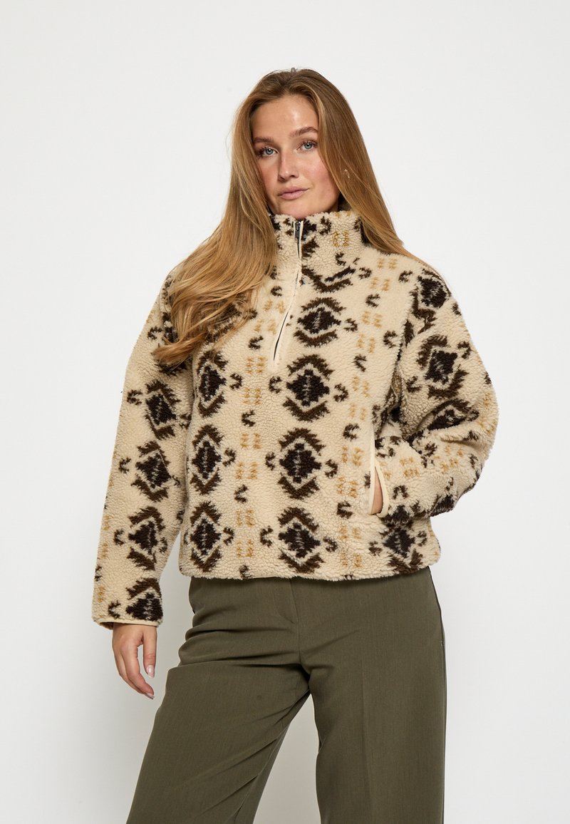 Béžový fleece sveter s čiernymi a hnedými geometrickými vzormi, so zipsovým golierom a bočnými vreckami. Teplá, textúrovaná látka.