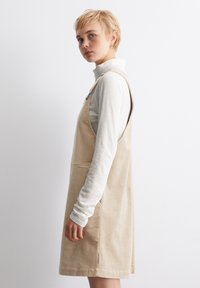 Béžové kordové šaty s nastavitelnými ramínky a přední kapsou, vrstvené přes lehký dlouhý rukáv. Jednoduchý, texturovaný design.