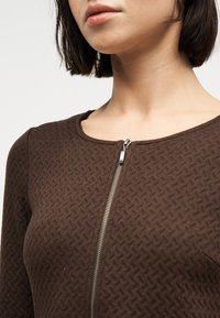 Γυναίκα με κοντά σκούρα μαλλιά που φοράει καφέ υφασμάτινο πουλόβερ με φερμουάρ και στρογγυλή λαιμόκοψη, εμφανιζόμενη από τους ώμους μέχρι το πηγούνι.