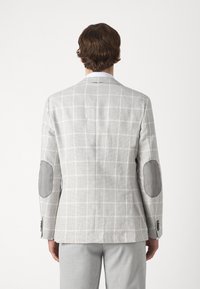Lichtgrijze geruite blazer van een gestructureerde stof, met contrasterende grijze elleboogpatches en een getailleerde snit. Opvallende knoopdetails op de mouwen.