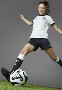 White and black sports jersey with short sleeves, black shorts, and black football boots; player kicking a black and white soccer ball on grass.