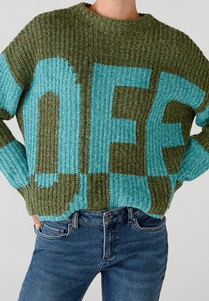 Personne portant un pull en grosse maille vert et bleu avec de grandes lettres en bloc et un jean bleu, les mains sur les hanches.