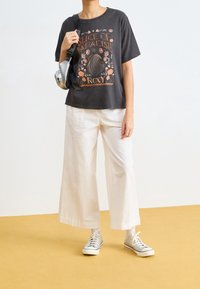 Šedé grafické tričko s designem "Kousek ráje", spárované s volnými bílými lněnými kalhotami a stříbrnými teniskami. Jednoduchý, ležérní outfit.
