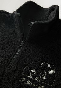 Schwarzer Fleece-Half-Zip-Pullover mit strukturiertem Obermaterial. Mit einem weißen bestickten Grafikmotiv eines Hundes und eines Vogels, darunter japanischer Text.
