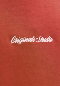 Czerwony materiał o gładkiej strukturze z białym haftowanym napisem "Originals Studio" w kursywie.