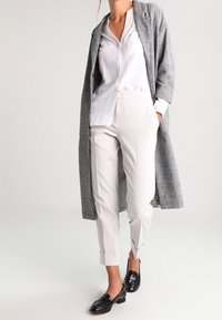 Lungo cappotto grigio a quadri sopra una camicia bianca con bottoni e pantaloni beige chiaro. Mocassini neri con finitura lucida completano l'outfit.