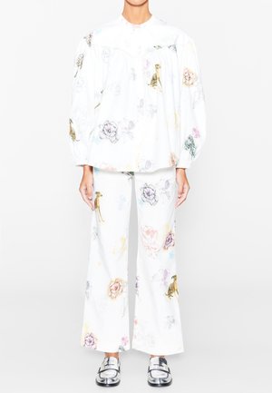 Kobieta w białej luźnej bluzce i szerokich spodniach z kolorowymi nadrukami psów i kwiatów, dobranych do srebrnych mokasynów, stojąca na białym tle.