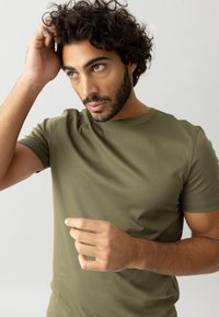 Olivgrünes kurzärmliges Baumwoll-T-Shirt mit klassischem Rundhalsausschnitt und glatter Textur. Das Model hat lockige Haare und gestikuliert leicht mit der Hand.