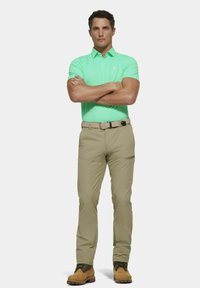 Mannen polo shirt in mintgroen, beige broek, elastische riem, bruine laarzen. Glad materiaal, slim-fit ontwerp, zichtbare zijzak op de broek.