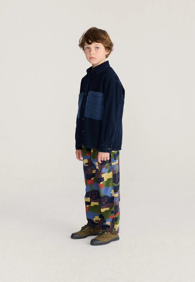 Garçon debout en pantalon camouflage multicolore, chemise bleu foncé à boutons avec grandes poches avant, et chaussures vert olive sur fond clair uni.
