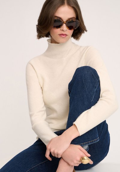 Pulover tricotat cu guler înalt de culoare crem, asortat cu blugi negri. Modelul poartă ochelari oversized cu ramă de broască țestoasă și un inel de aur, într-o poziție aşezată.